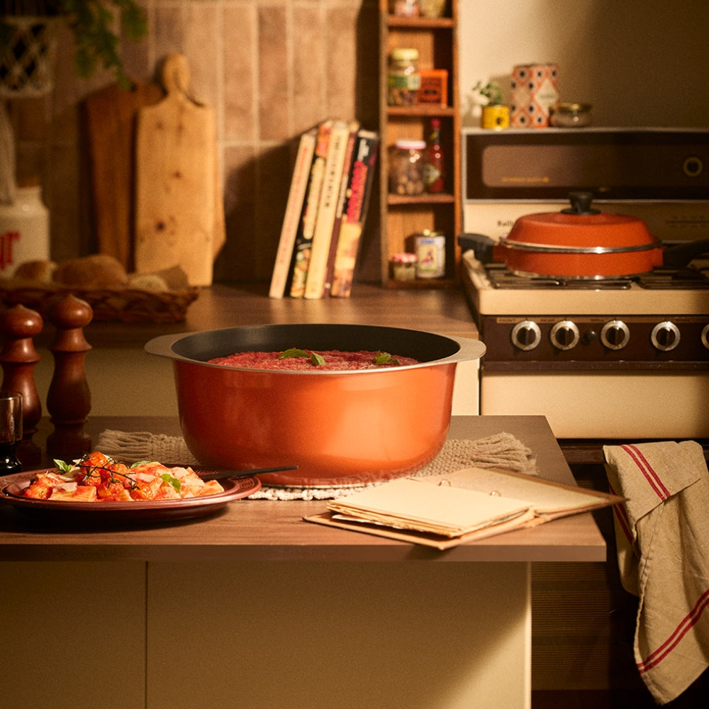 An orange Bessemer with a black interior sits on a kitchen counter, filled with a red stew or soup. The background shows a warm, cozy kitchen setting with a stove, books, and wooden cutting boards.