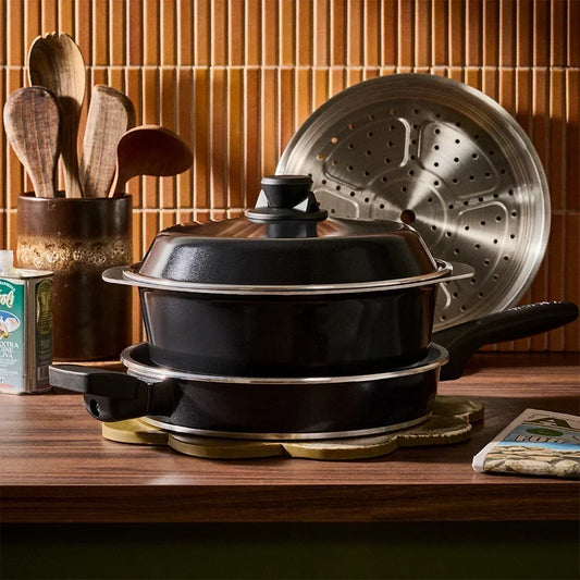 A covered black pot rests inside a frying pan on a wooden countertop, and a metal steamer insert is positioned in the background next to a container of wooden utensils