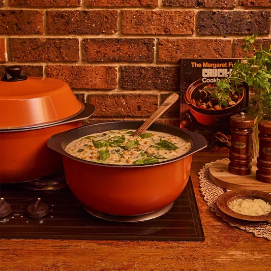 An orange pot filled with creamy soup is placed on a stovetop against an exposed brick wall, accompanied by a cookbook, wooden salt and pepper shakers, and fresh herbs.