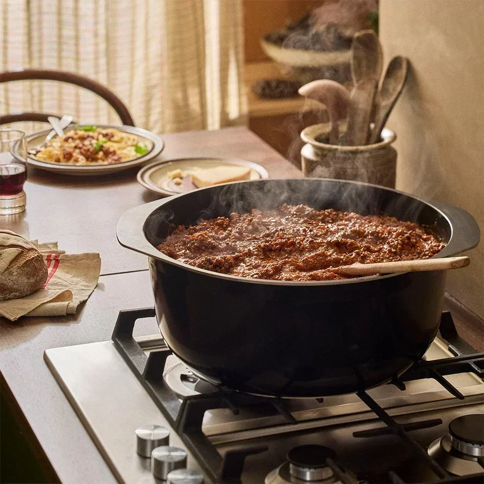 A large black pot sits on a stovetop, and steaming chili simmers inside the pot with a wooden spoon resting across the rim.