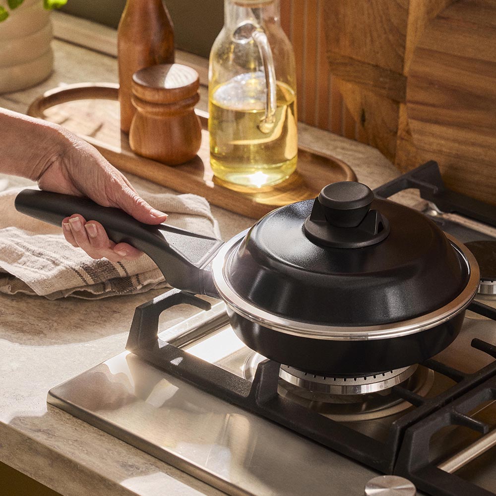 Black frying pan with lid on a gas stove, hand holding handle, wooden kitchen counter in background