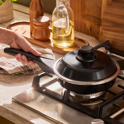 Black frying pan with lid on a gas stove, hand holding handle, wooden kitchen counter in background
