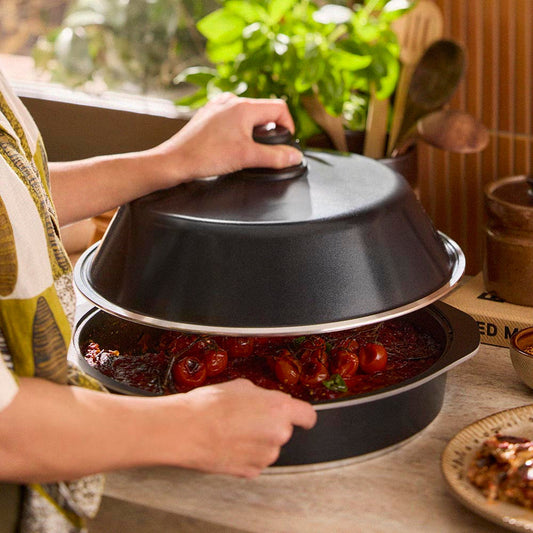 Person holding a black lid over a dish of food on a wooden table with plants in the background