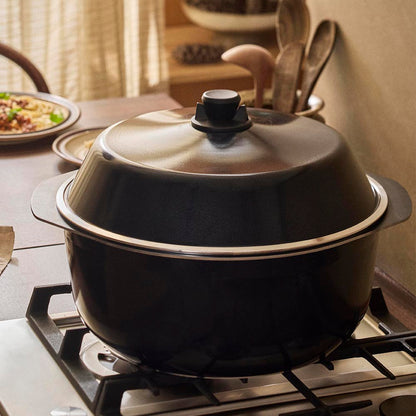 Black pot with a lid on a stove, surrounded by kitchen utensils and food.