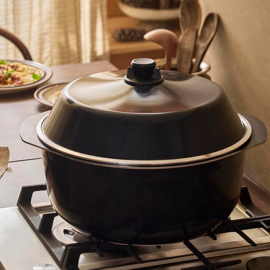 Black pot with a lid on a stove, surrounded by kitchen utensils and food.