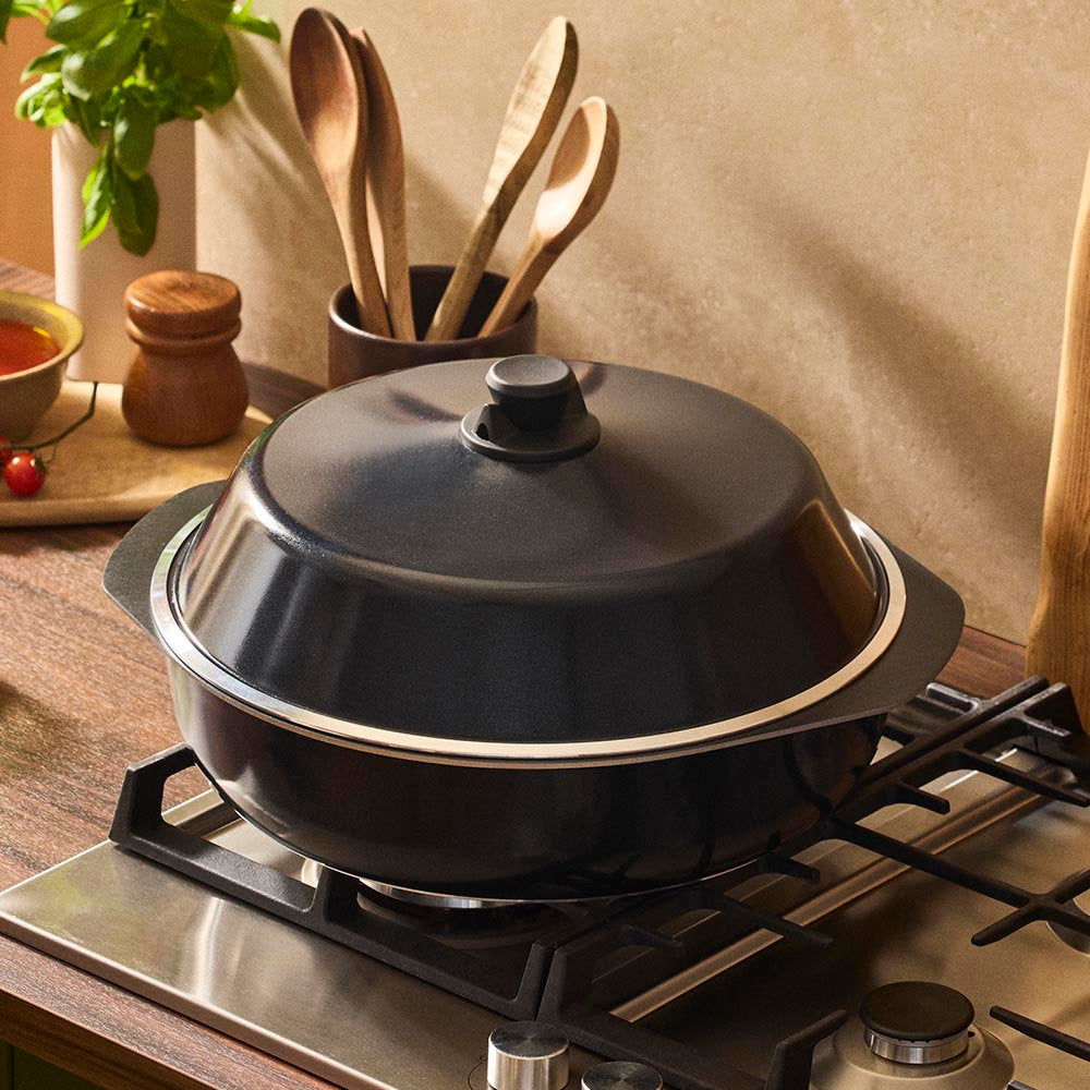 Black pot with a lid on a stove, surrounded by wooden utensils and ingredients.