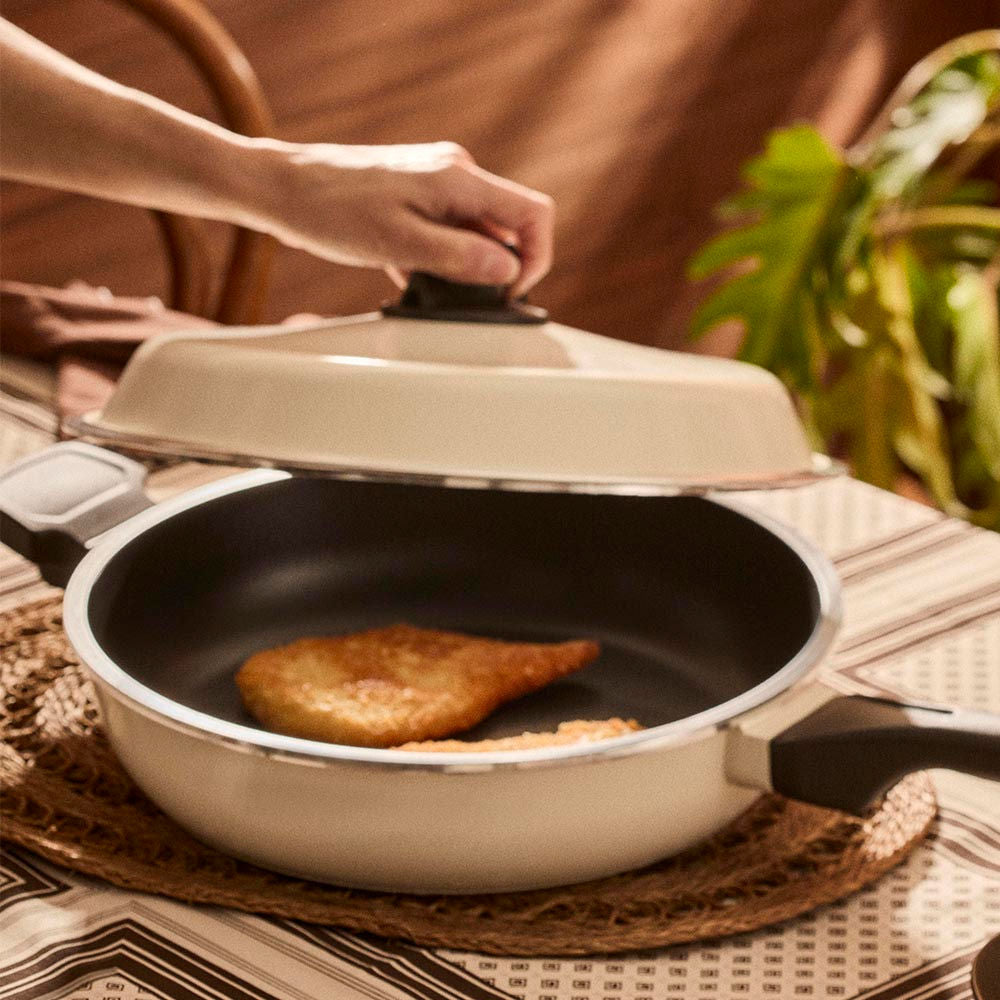 Person cooking food in a frying pan with a lid on a table.