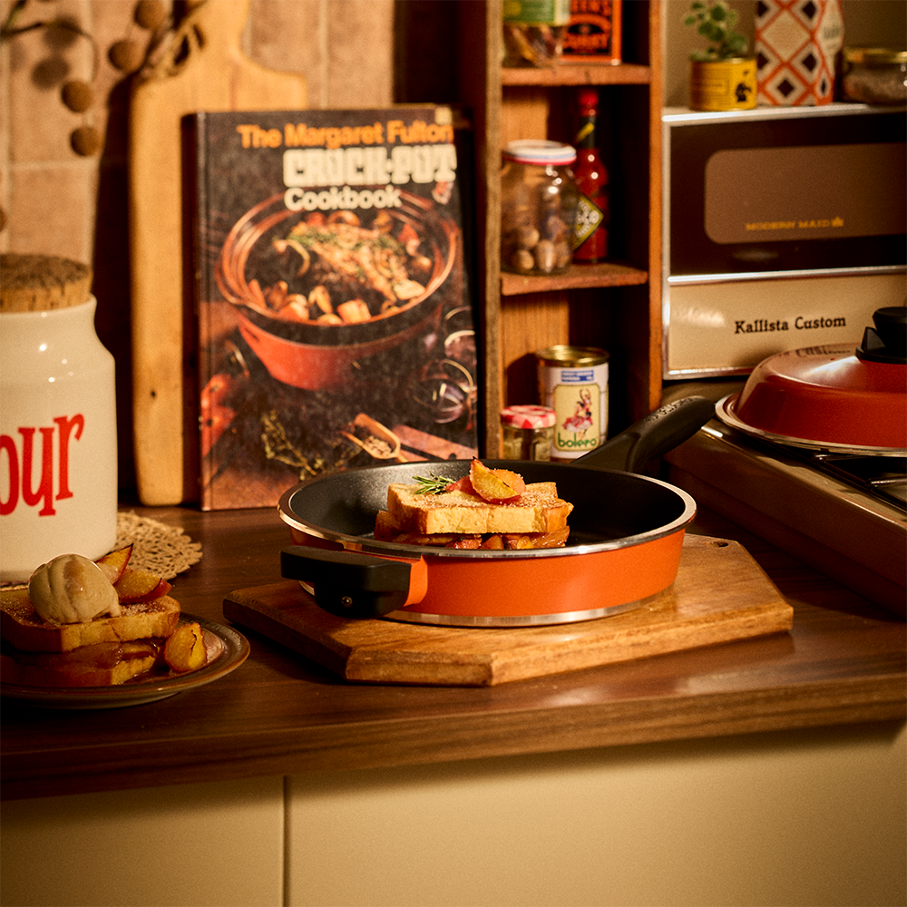 A kitchen countertop scene with a frying pan holding toasted bread, a cookbook titled 'The Margaret Fulton Chick-Put Cookbook' standing behind it, and warm-toned kitchen appliances and shelves in the background.