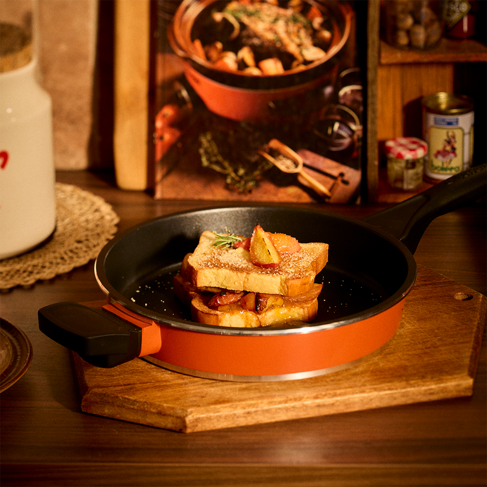 An orange and black Bessemer nonstick frying pan sits on a wooden cutting board on a dark wooden table, filled with a stack of toasted bread slices. The background is a warm, rustic kitchen setting with a cookbook and jars.