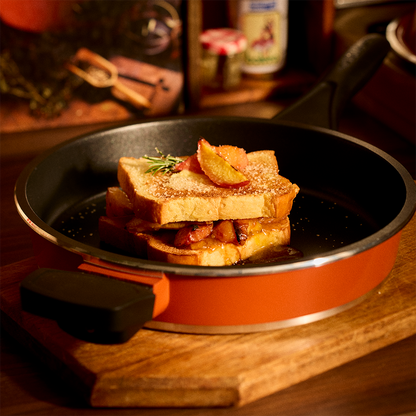 An orange and black Bessemer frying pan on a wooden board, holding a stack of French toast with bacon and a slice of apple on a warm wooden table.