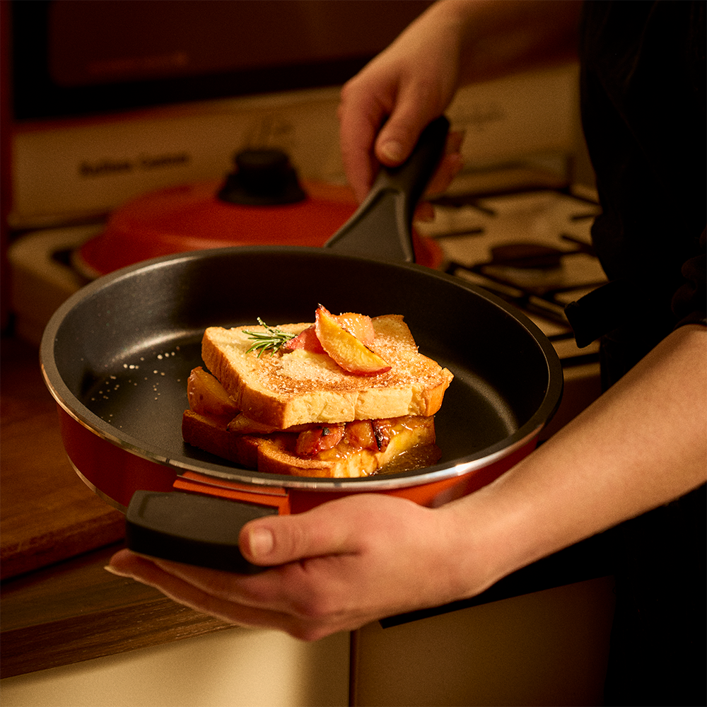 A person holds an orange Bessemer non-stick frying pan with a black handle, cooking a stack of French toast slices with a strawberry sauce and rosemary garnish in a warm kitchen setting.