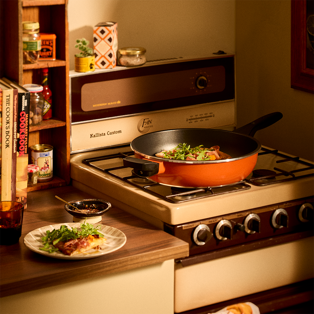 A retro-style kitchen scene with a vintage cream and brown gas stove. An orange Bessemer frying pan sits on the front burner, cooking vegetables, while a small plate of food rests on the counter beside it.