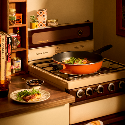 A retro-style kitchen scene with a vintage cream and brown gas stove. An orange Bessemer frying pan sits on the front burner, cooking vegetables, while a small plate of food rests on the counter beside it.