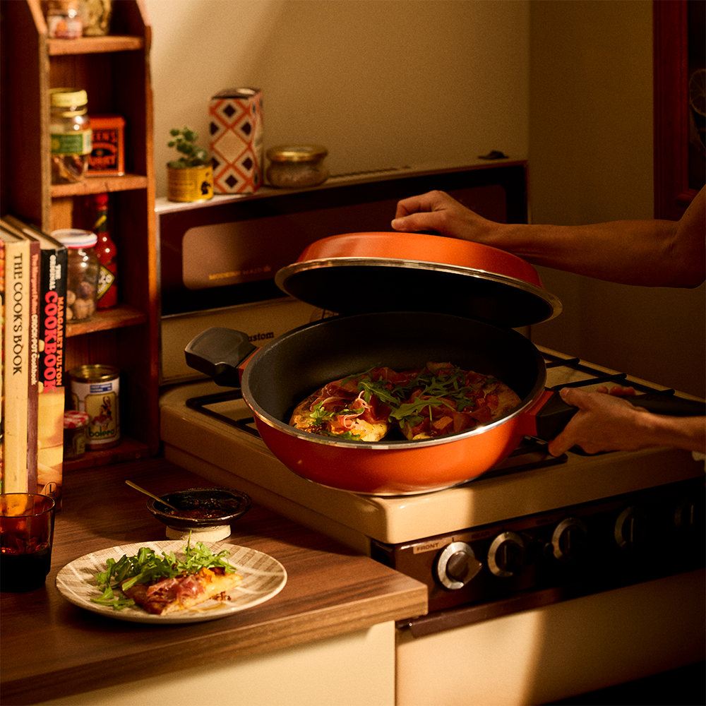 An orange nonstick Bessemer frying pan with a lid is being lifted by a person on a cream gas stove in a warm, dimly lit kitchen. A plate of food sits on the wooden countertop nearby.