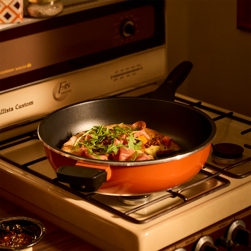 An orange Bessemer non-stick frying pan with a black handle sits on a lit gas stove burner, cooking food with green herbs. The background shows a cream-colored stove and a warm, cozy kitchen environment.
