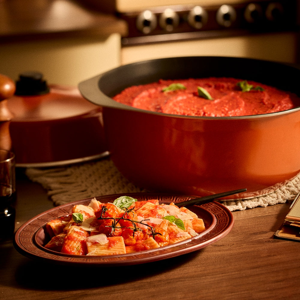 A large orange Bessemer pot filled with red sauce, shown alongside a plated pasta dish on a wooden table in a warm kitchen setting.