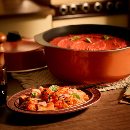 A large orange Bessemer pot filled with red sauce, shown alongside a plated pasta dish on a wooden table in a warm kitchen setting.