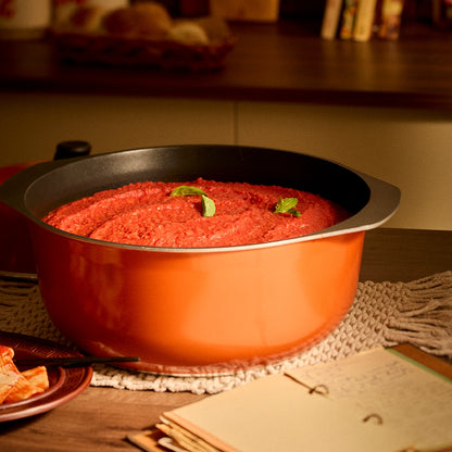 A large orange Bessemer casserole with a black interior, filled with a red tomato-based sauce and garnished with green basil leaves, set on a woven placemat on a wooden table.