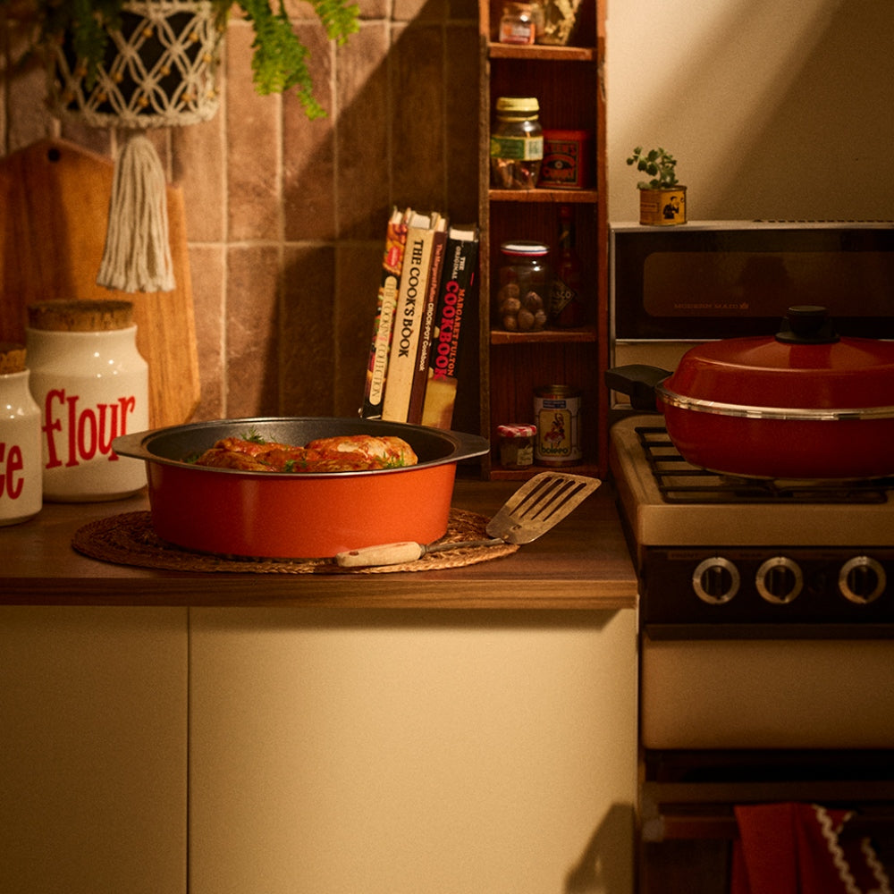 A warm, cozy kitchen scene with an orange dutch oven containing cooked food on a wooden countertop, next to a matching red pot on a stove. The background features a tiled wall, wooden shelves with cookbooks and jars, and a microwave.