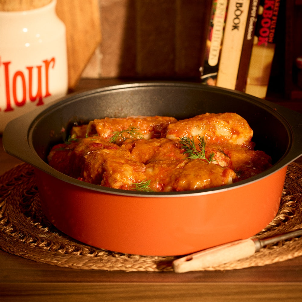 An orange Bessemer dutch oven filled with chicken and dumplings in a rich tomato sauce, set on a woven placemat with a wooden spoon handle resting beside it.
