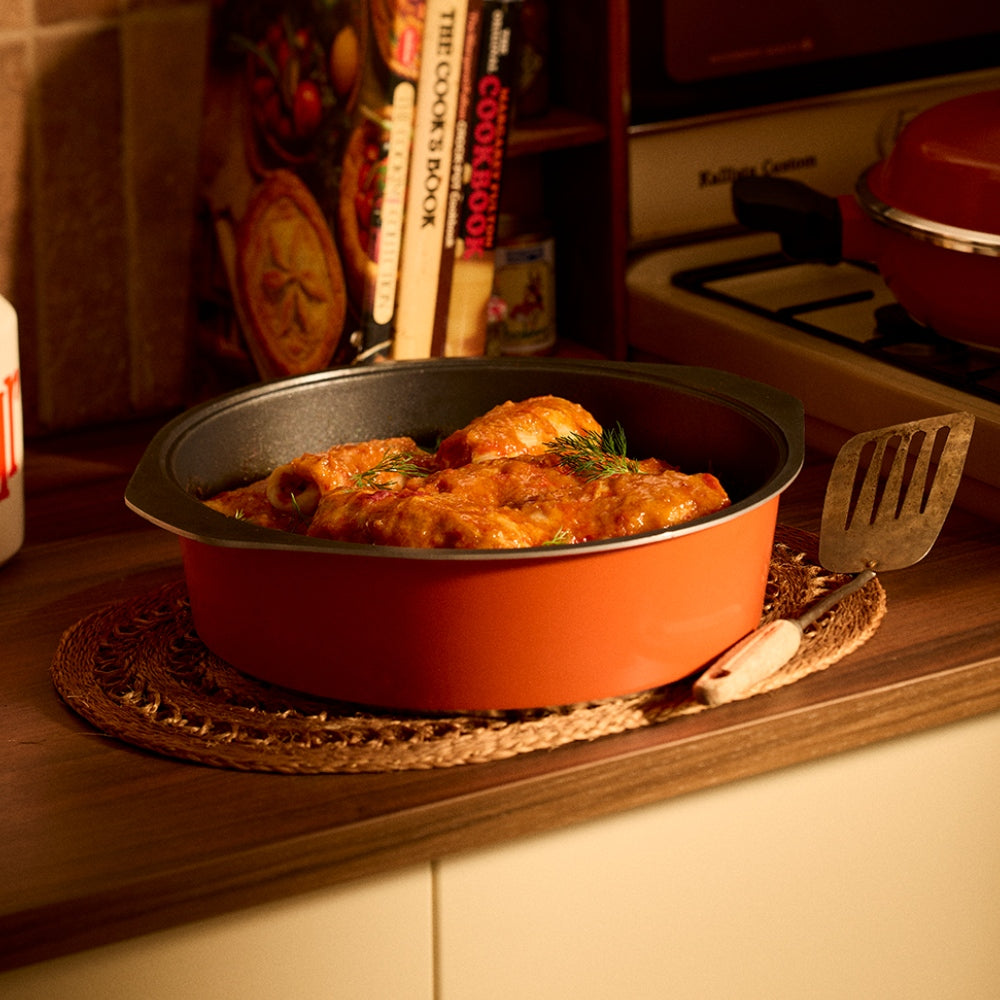 An orange nonstick Bessemer dutch oven filled with pasta and sauce, sitting on a woven placemat on a wooden kitchen counter with cookbooks and a stove in the background.