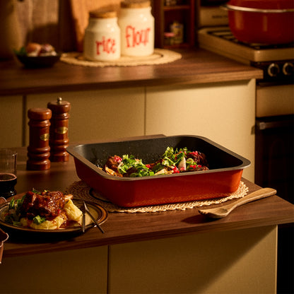 A rectangular orange Bessemer non-stick baking pan filled with cooked food, set on a wooden kitchen countertop with a plate of food, salt and pepper mills, and kitchen canisters in the background.