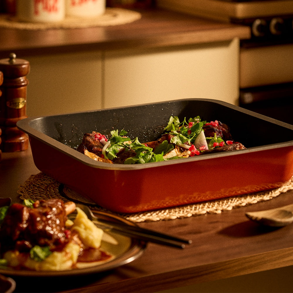 A rectangular non-stick roasting pan with an orange exterior and black interior, shown filled with cooked meat and green herbs on a wooden table in a warm kitchen setting.