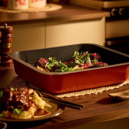 A rectangular non-stick roasting pan with an orange exterior and black interior, shown filled with cooked meat and green herbs on a wooden table in a warm kitchen setting.