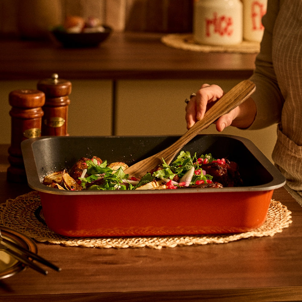 A person in a kitchen uses a wooden spoon to stir a colorful salad inside a rectangular orange non-stick Bessemer baking pan, set on a woven placemat over a wooden table.