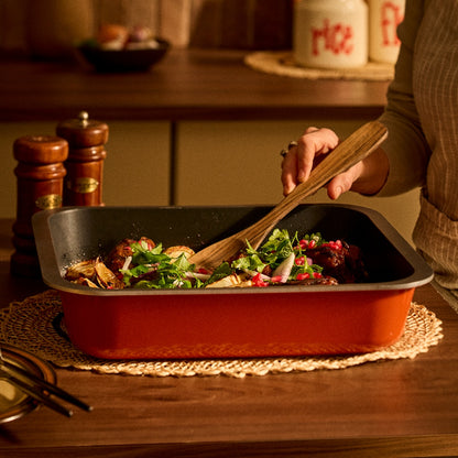A person in a kitchen uses a wooden spoon to stir a colorful salad inside a rectangular orange non-stick Bessemer baking pan, set on a woven placemat over a wooden table.