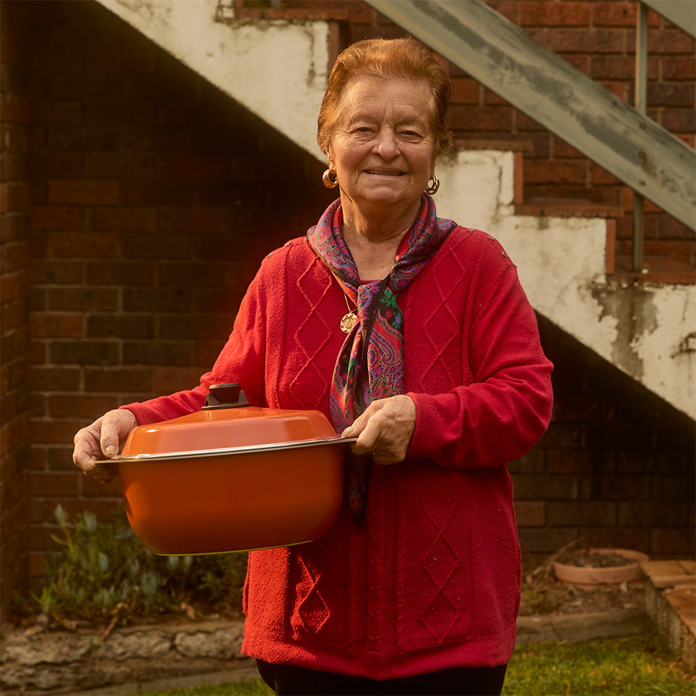An older woman stands outdoors in front of a brick wall and staircase, holding a large orange Bessemer casserole dish with a lid.