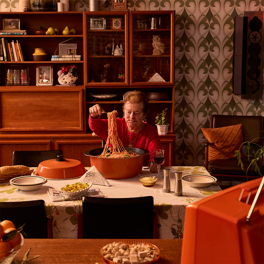 A woman in a red sweater sits at a dining table holding an orange Bessemer pot filled with spaghetti in a warm, retro-styled room with wood cabinets and patterned wallpaper.