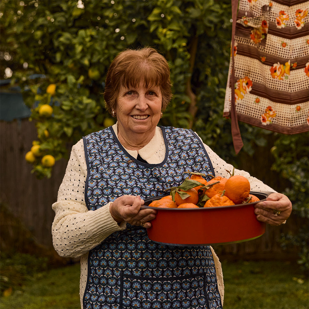 An older woman smiling in a garden while holding a orange Bessemer dutch oven filled with oranges. She is wearing a cream knit sweater under a blue floral apron, with a crocheted garment hanging in the background.