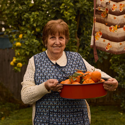 An older woman smiling in a garden while holding a orange Bessemer dutch oven filled with oranges. She is wearing a cream knit sweater under a blue floral apron, with a crocheted garment hanging in the background.