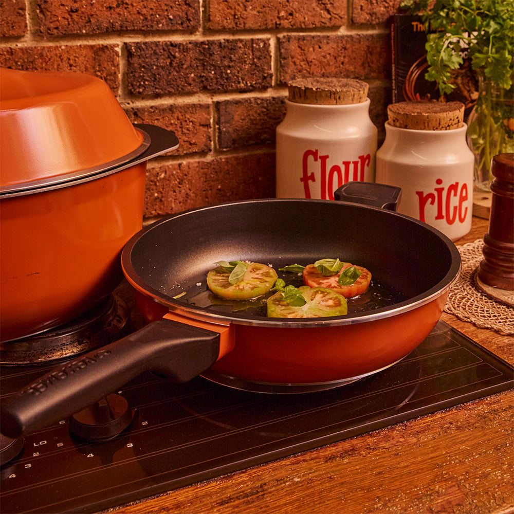 A non-stick Bessemer frying pan with a burnt orange exterior and black handle sits on a stovetop, cooking sliced tomatoes. The background features a brick wall, kitchen canisters labeled flour and rice, and a potted plant.