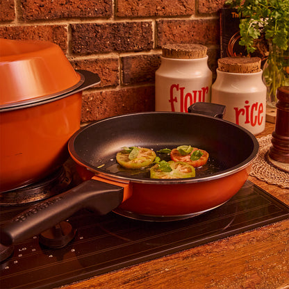 A non-stick Bessemer frying pan with a burnt orange exterior and black handle sits on a stovetop, cooking sliced tomatoes. The background features a brick wall, kitchen canisters labeled flour and rice, and a potted plant.