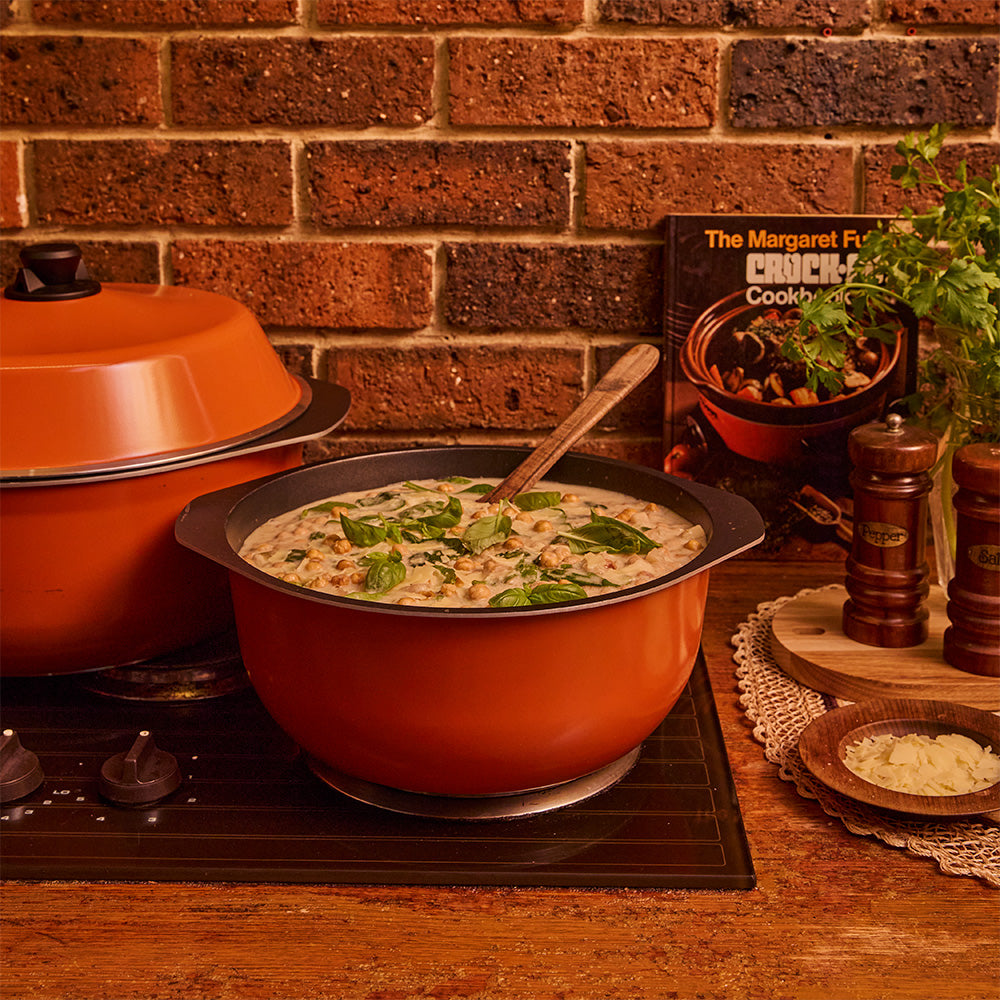 An orange Bessemer casserole filled with a creamy mushroom soup sits on a stovetop in a kitchen scene with a brick wall background, a second pot behind it, and cookbooks and seasoning bottles nearby.