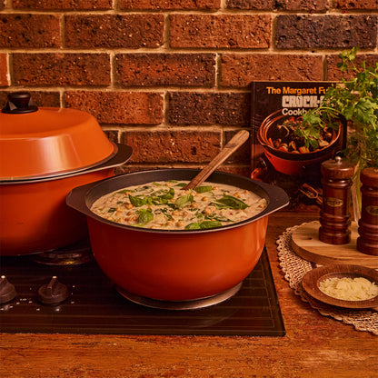 An orange Bessemer casserole filled with a creamy mushroom soup sits on a stovetop in a kitchen scene with a brick wall background, a second pot behind it, and cookbooks and seasoning bottles nearby.