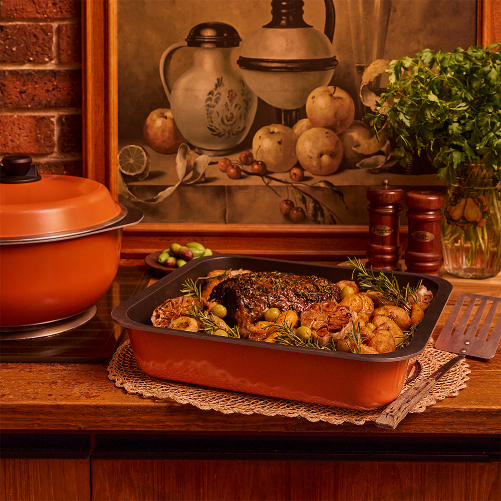 An orange Bessemer ceramic baking dish filled with roasted meat, potatoes, and rosemary sits on a wooden countertop in a warm kitchen setting, with a matching orange pot, salt and pepper shakers, and a decorative painting in the background.
