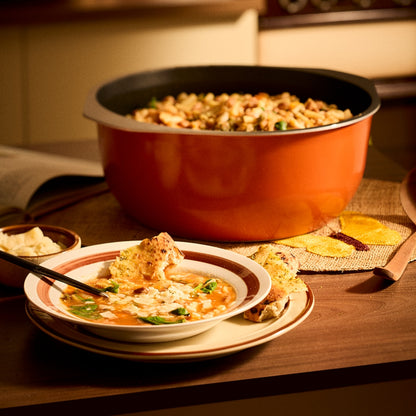 An orange Bessemer Dutch oven filled with stuffing, set on a wooden table next to a bowl of soup and bread in a warm, cozy kitchen setting.