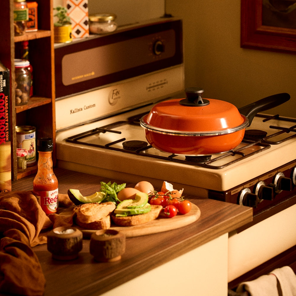 An orange Bessemer frying pan with a matching lid sits on a retro-style cream and brown gas stove in a warm, cozy kitchen setting. A wooden cutting board with avocado, tomatoes, eggs, and toast is prepared on the counter in the foreground.