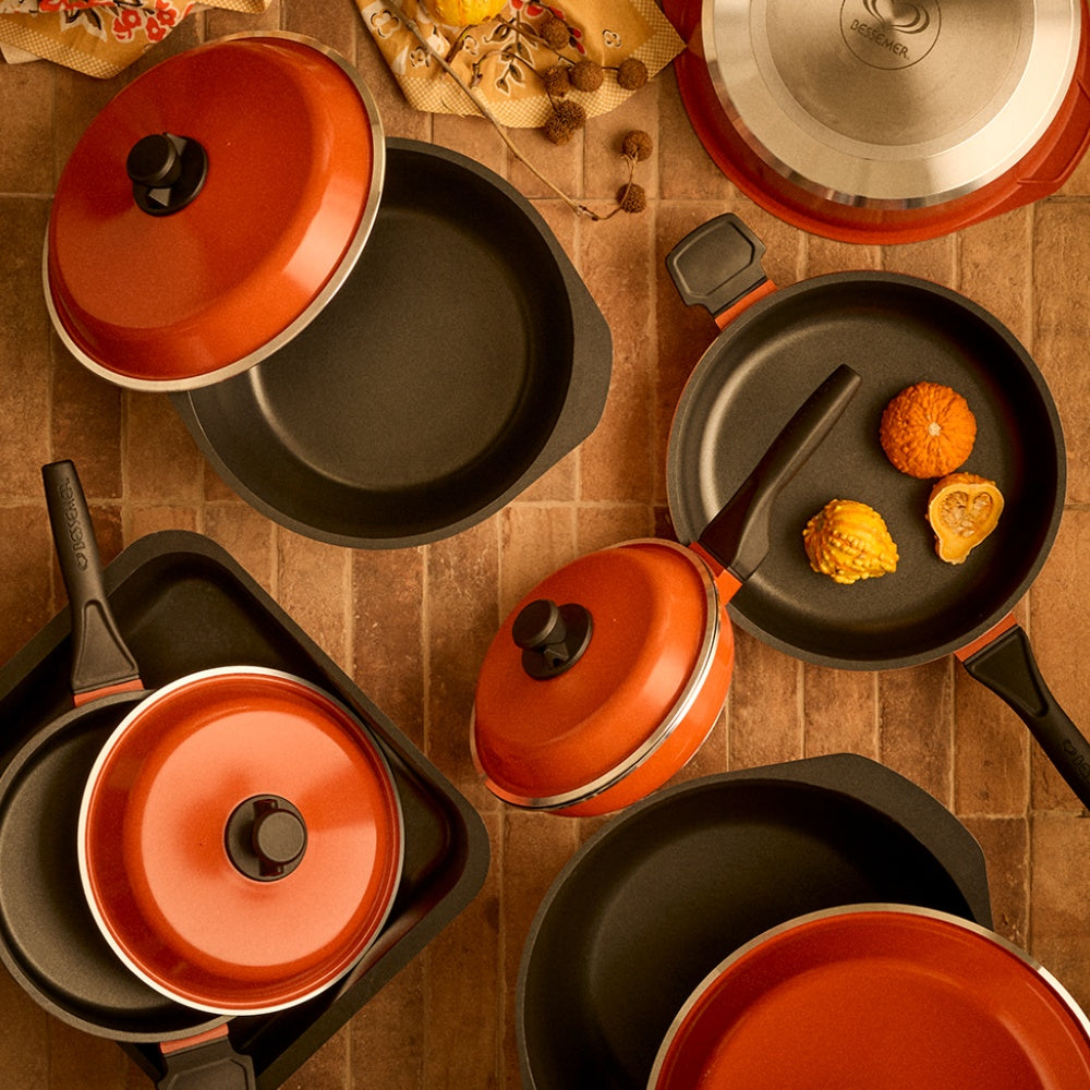 A flat-lay arrangement of a Bessemer cookware set with orange lids and black non-stick pans on a brown tiled surface. Several pans are shown empty, while one pan contains small orange food pieces for scale.